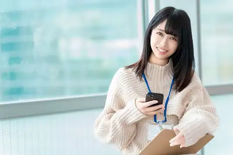 オフィスの窓辺でスマートフォンとバインダーを持つスタッフの笑顔